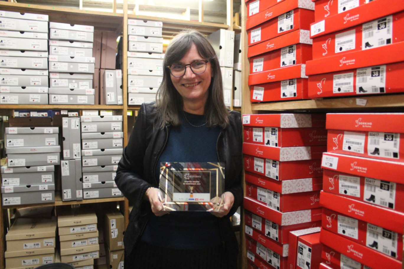 Sylvie Laboue de la boutique Un pas de Plus à Soissons pose au sein de sa réserve avec le trophée Orange du numérique décerné par la CCI et récompensant la création de son site Internet de déstockage de chaussures.